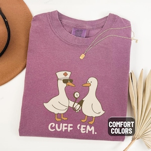 May include: Berry-colored t-shirt with a graphic of two cartoon geese, one wearing a nurse's hat and sunglasses, holding a thermometer. The text "CUFF 'EM" is below the geese. The shirt has a Comfort Colors tag.