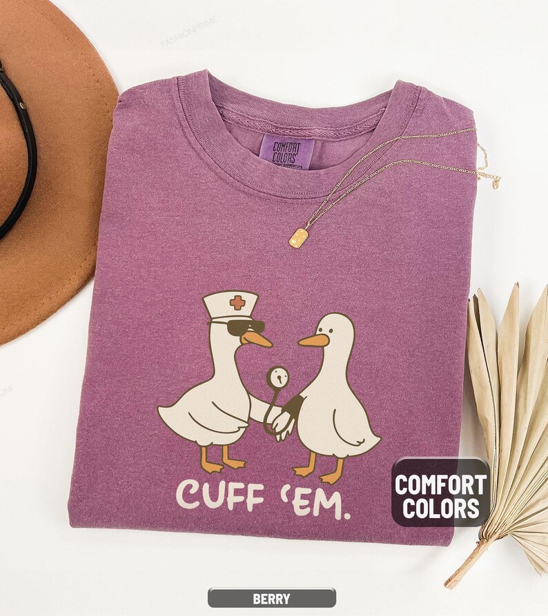 May include: Berry-colored t-shirt with a graphic of two cartoon geese, one wearing a nurse's hat and sunglasses, holding a thermometer. The text "CUFF 'EM" is below the geese. The shirt has a Comfort Colors tag.