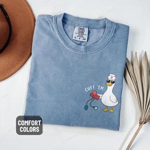 May include: A blue jean Comfort Colors t-shirt with a cartoon duck wearing sunglasses and a nurse's hat, holding a blood pressure cuff. The text "CUFF 'EM" is above the duck.