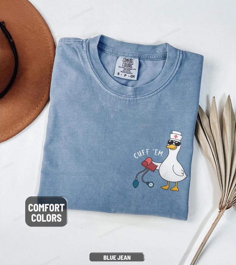 May include: A blue jean Comfort Colors t-shirt with a cartoon duck wearing sunglasses and a nurse's hat, holding a blood pressure cuff. The text "CUFF 'EM" is above the duck.