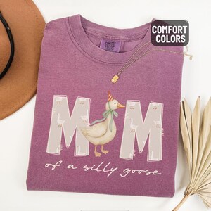 May include: Berry-colored t-shirt with the words "MOM of a silly goose" and a graphic of a goose wearing a party hat and bow. The shirt is made of a soft material and has a crew neck. The Comfort Colors brand tag is visible.