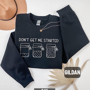 May include: Black crewneck sweatshirt with the text "DON'T GET ME STARTED" and three illustrated jars. The sweatshirt has long sleeves and a round neckline. The brand name "GILDAN" is visible on the tag.