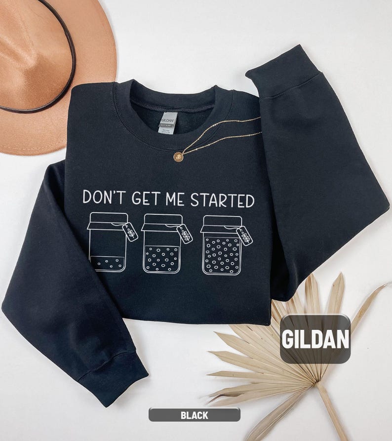 May include: Black crewneck sweatshirt with the text "DON'T GET ME STARTED" and three illustrated jars. The sweatshirt has long sleeves and a round neckline. The brand name "GILDAN" is visible on the tag.