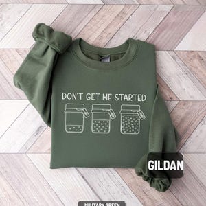 May include: Military green sweatshirt with the text "DON'T GET ME STARTED" and three jar illustrations. The sweatshirt is made of a soft material and is a comfortable fit. The brand name "GILDAN" is visible.