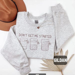 May include: A light gray sweatshirt with the text "DON'T GET ME STARTED" and three jar illustrations. The sweatshirt is made by Gildan and is labeled "ASH". A brown hat and a necklace are also visible.