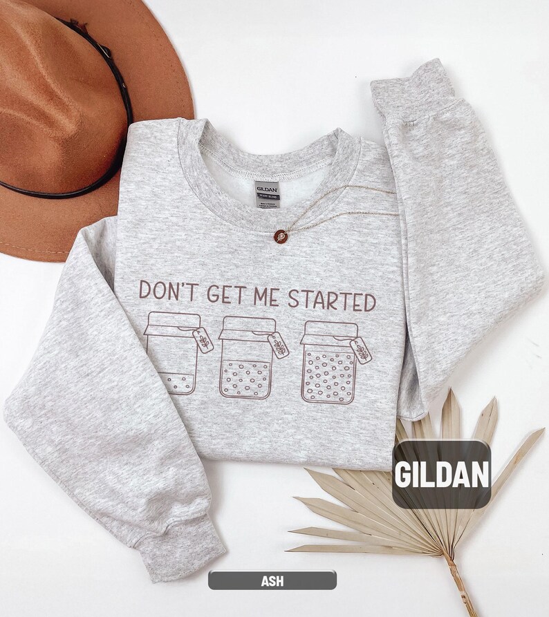 May include: A light gray sweatshirt with the text "DON'T GET ME STARTED" and three jar illustrations. The sweatshirt is made by Gildan and is labeled "ASH". A brown hat and a necklace are also visible.