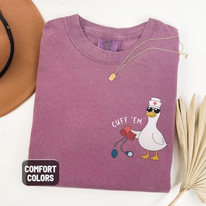 May include: Berry-colored Comfort Colors t-shirt with a cartoon goose wearing sunglasses and a nurse's hat, holding a blood pressure cuff. The text "CUFF 'EM" is above the goose. A gold necklace is draped across the shirt.