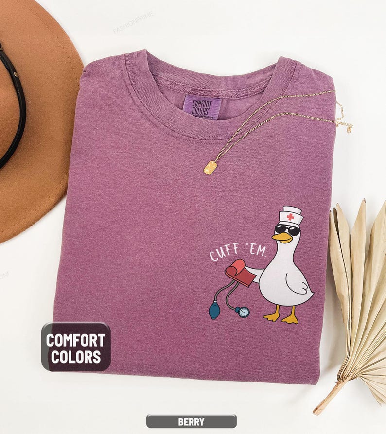 May include: Berry-colored Comfort Colors t-shirt with a cartoon goose wearing sunglasses and a nurse's hat, holding a blood pressure cuff. The text "CUFF 'EM" is above the goose. A gold necklace is draped across the shirt.