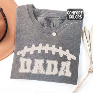 Puede incluir: Una camiseta gris Comfort Colors con un gr&aacute;fico de f&uacute;tbol desgastado y la palabra "DADA" en una fuente de bloque. La camiseta est&aacute; doblada y tiene una peque&ntilde;a etiqueta en el cuello. Un collar con un colgante redondo est&aacute; sobre la camiseta. La camiseta est&aacute; etiquetada como "Pepper".