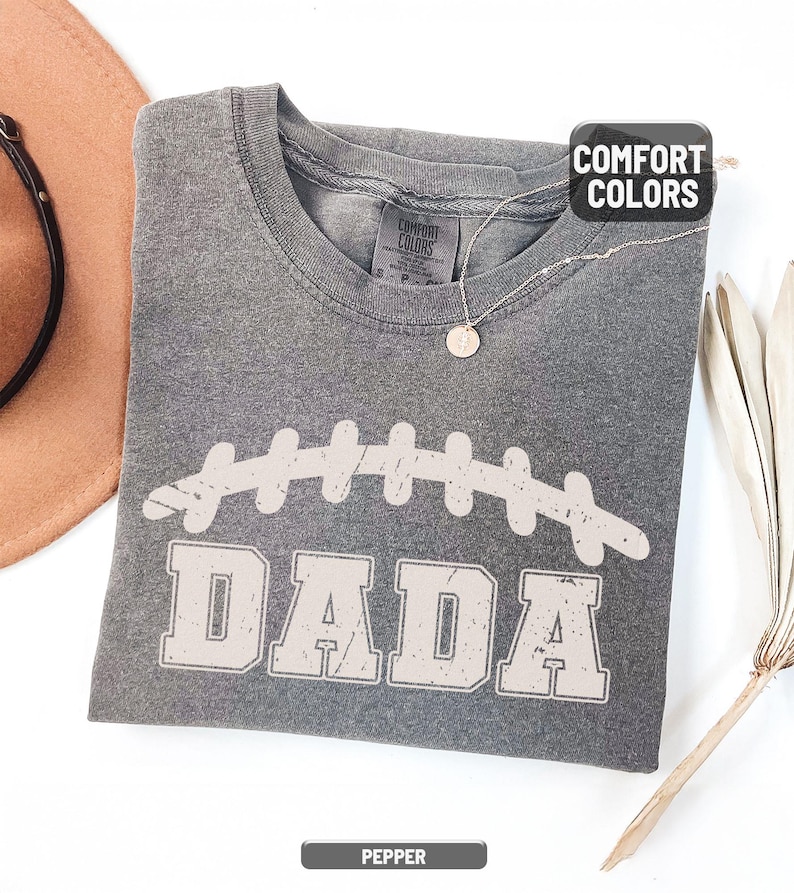 Puede incluir: Una camiseta gris Comfort Colors con un gr&aacute;fico de f&uacute;tbol desgastado y la palabra "DADA" en una fuente de bloque. La camiseta est&aacute; doblada y tiene una peque&ntilde;a etiqueta en el cuello. Un collar con un colgante redondo est&aacute; sobre la camiseta. La camiseta est&aacute; etiquetada como "Pepper".