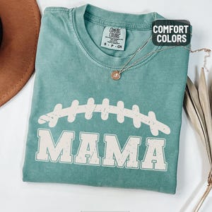 Puede incluir: Camiseta verde claro Comfort Colors con un gr&aacute;fico de f&uacute;tbol americano y la palabra "MAMA" en letras blancas. Un collar de oro con un colgante est&aacute; drapeado sobre la camiseta. La camiseta est&aacute; doblada sobre una superficie blanca.