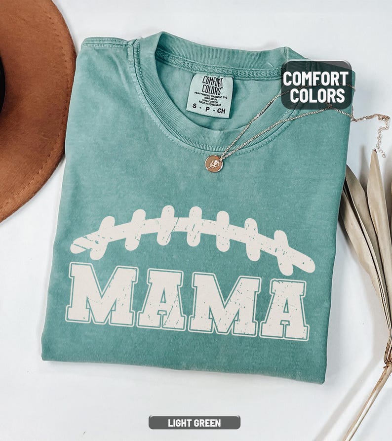 Puede incluir: Camiseta verde claro Comfort Colors con un gr&aacute;fico de f&uacute;tbol americano y la palabra "MAMA" en letras blancas. Un collar de oro con un colgante est&aacute; drapeado sobre la camiseta. La camiseta est&aacute; doblada sobre una superficie blanca.