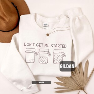 May include: White sweatshirt with the phrase "DON'T GET ME STARTED" printed in dark gray. The design includes three jars with varying contents and tags. The brand name "GILDAN" is visible. A small gold necklace is also present.