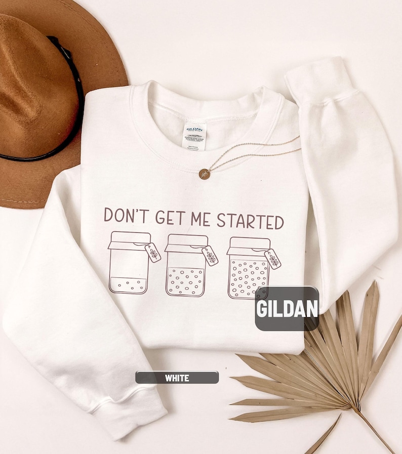 May include: White sweatshirt with the phrase "DON'T GET ME STARTED" printed in dark gray. The design includes three jars with varying contents and tags. The brand name "GILDAN" is visible. A small gold necklace is also present.