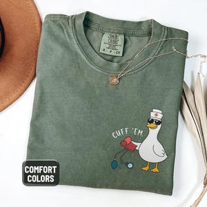 May include: Moss green Comfort Colors t-shirt with a cartoon duck wearing sunglasses and a nurse's hat, holding a blood pressure cuff. The text "CUFF 'EM" is above the duck. A gold necklace is draped on the shirt.