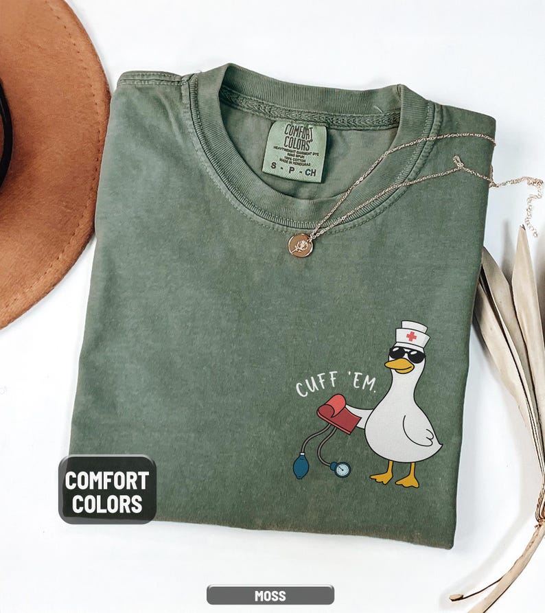May include: Moss green Comfort Colors t-shirt with a cartoon duck wearing sunglasses and a nurse's hat, holding a blood pressure cuff. The text "CUFF 'EM" is above the duck. A gold necklace is draped on the shirt.