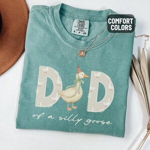May include: Light green Comfort Colors t-shirt with the word "DAD" in large letters, each letter decorated with a goose pattern. A cartoon goose wearing a party hat is in the center. The text "of a silly goose" is below.