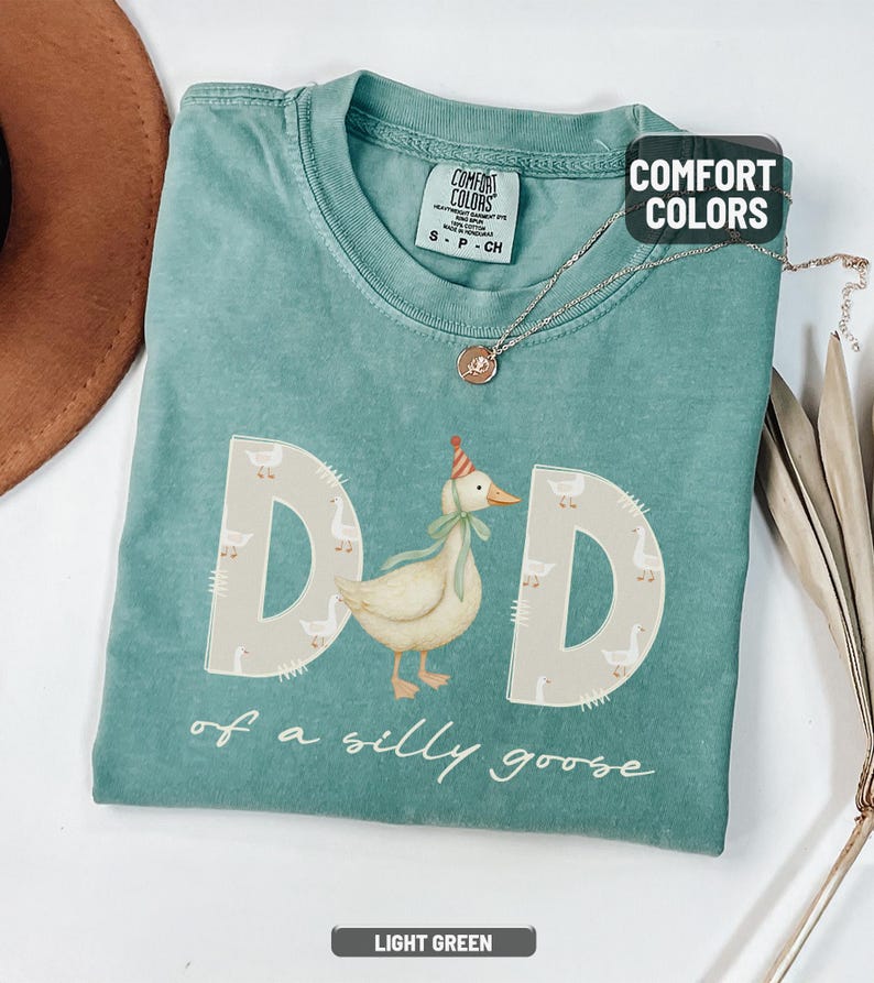 May include: Light green Comfort Colors t-shirt with the word "DAD" in large letters, each letter decorated with a goose pattern. A cartoon goose wearing a party hat is in the center. The text "of a silly goose" is below.