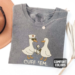 May include: Pepper-colored Comfort Colors t-shirt featuring a cartoon design of two geese. One goose is wearing a nurse's hat and sunglasses, holding hands with another goose. The text "CUFF 'EM" is printed below the geese.