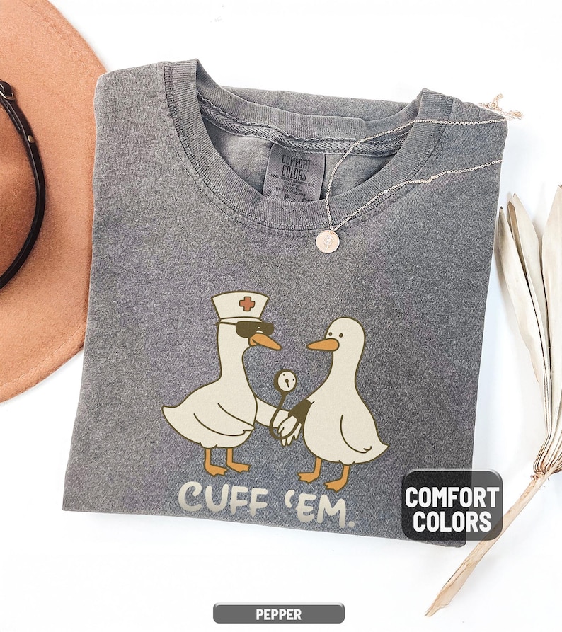 May include: Pepper-colored Comfort Colors t-shirt featuring a cartoon design of two geese. One goose is wearing a nurse's hat and sunglasses, holding hands with another goose. The text "CUFF 'EM" is printed below the geese.