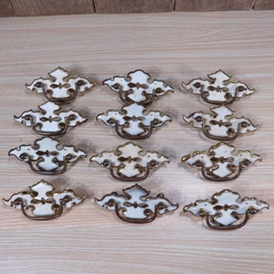 Peut inclure: Douze tirettes de tiroir de cabinet antiques en blanc et or avec des motifs ornés. Chaque tirette a une poignée en boucle.