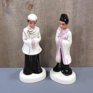 May include: Pair of ceramic figurines depicting Asian figures. The figurines are dressed in white robes with pink and gold accents, and black pants. The figures stand on white oval bases. One figure wears a white hat with pink trim.
