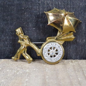 May include: Gold-tone brooch featuring a rickshaw with a driver and a passenger, with a gold-tone umbrella on top. The rickshaw wheel is decorated with rhinestones.