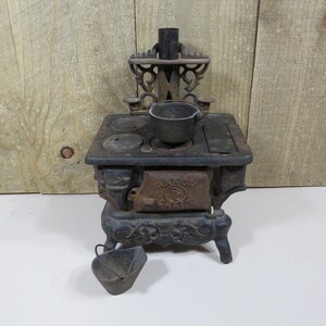 May include: A miniature black cast iron stove featuring ornate scrollwork, a chimney, and a front door embossed with the text RANCH OAK. The stove includes a small removable pot on the stovetop and a separate matching bucket placed in front.