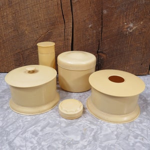 May include: Set of five vintage cream-colored vanity jars with lids. The jars are made of a smooth, hard plastic and have a slightly rounded shape. The lids are all different shapes and sizes, and some have a decorative design.