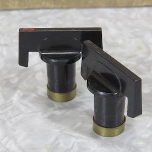 May include: Two black knobs with a rectangular handle and a cylindrical base. The base has a gold-coloured ring. One knob has a small red accent on the handle. These knobs are likely for a piece of vintage equipment.