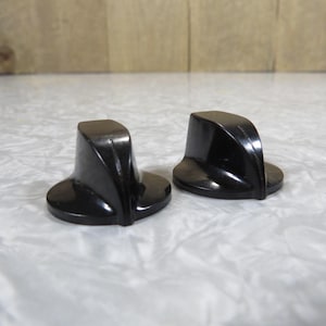 May include: Two black, vintage-style knobs with a rounded base and a curved top. The knobs are made of a glossy, dark material and are likely for a stove or appliance. The knobs are set against a light-colored surface.