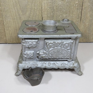 May include: A vintage cast iron toy stove in a silver finish with ornate floral relief detailing. The front door features the embossed text EAGLE. A small metal pot sits on the stovetop, and a dark metal bucket is placed in front.