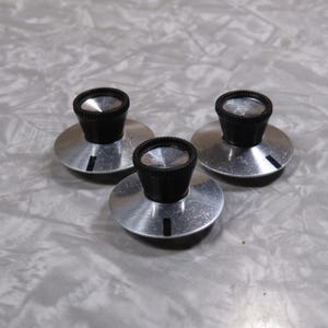 May include: Three silver-colored knobs with black tops. Each knob has a circular base and a cylindrical black top with a ridged texture. The knobs are arranged in a triangular pattern against a light gray background.