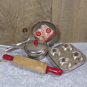 May include: Vintage toy kitchen set with a silver saucepan, lid, and frying pan, a wooden rolling pin with red handles, and a silver muffin tin. The frying pan has a label that reads "A Toy - One Egg -  Manufactured by The Toy Tinkers, Inc. - Made in U.S.A."