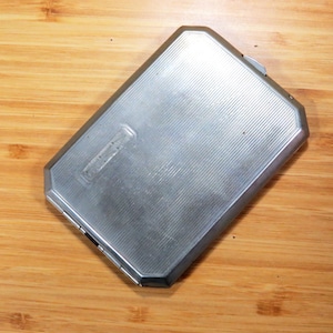 May include: A rectangular, silver-colored cigarette case with a ridged design. The case has a hinged lid and a small, rectangular emblem on the front. The case is made of metal and has a brushed finish.