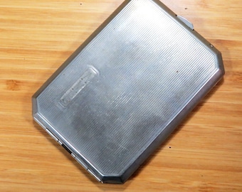 May include: A rectangular, silver-colored cigarette case with a ridged design. The case has a hinged lid and a small, rectangular emblem on the front. The case is made of metal and has a brushed finish.