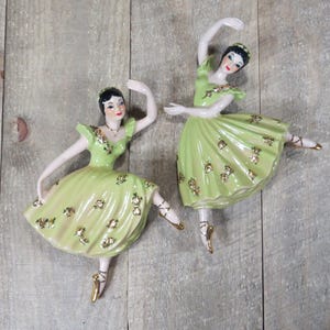 May include: Two ceramic ballerina figurines in graceful poses. Each figurine wears a light green dress adorned with gold accents and a matching headpiece. The ballerinas have dark hair and are posed on a wooden surface.