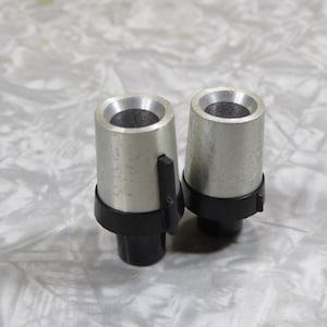 May include: Two silver-colored metal objects with a black base and a small black lever. The objects have a cylindrical shape with a hollow top. The background is a light gray, textured surface.