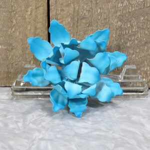May include: A light blue metal flower sculpture with layered petals. The flower is mounted on a clear glass block. The background features a weathered wooden surface.