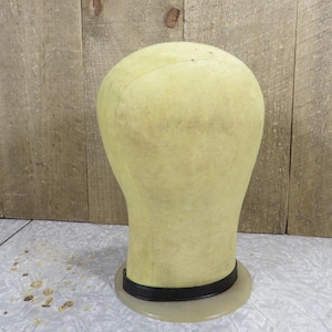 May include: A vintage, light yellow wig head form with a black band around the base. The head form is made of fabric and has a rounded top. The base is clear plastic. The background is a wooden wall.