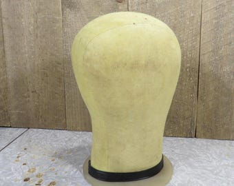 May include: A vintage, pale yellow wig head form with a black band around the base. The head form is made of fabric and has a rounded top. The base is clear plastic. The background is a wooden wall.