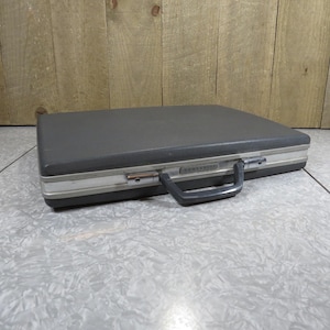 May include: A gray Samsonite briefcase with a silver trim and a black handle. The briefcase is rectangular and has a metal clasp. The background is a light gray and white patterned surface with a wooden wall.