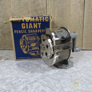 May include: A vintage Giant pencil sharpener with a silver finish and a gray base, next to its original blue and tan box. The box reads "AUTOMATIC GIANT PENCIL SHARPENER". The sharpener has a hand crank.