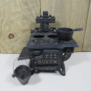 May include: A miniature black cast iron stove featuring the word QUEEN embossed on the front door. The stove includes a small matching pot resting on the top surface and a separate small bucket with a wire handle placed in front.