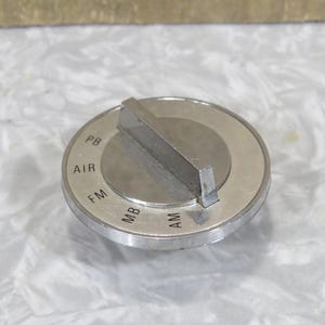 May include: A silver radio dial knob with a raised rectangular handle. The dial is marked with "PB", "AIR", "FM", "MB", and "AM" in a circle. The knob has a brushed metal finish and is likely for a vintage radio.