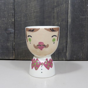 May include: A ceramic egg cup shaped like a person's head and torso. The figure features a tan face with green eyes, brown hair, and a brown mustache. The base is white with a pink bow tie featuring green polka dots and two small black buttons.