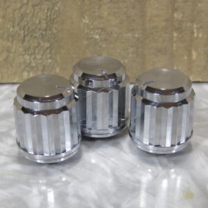 May include: Three shiny, chrome-plated control knobs. Each knob has a flat, circular top with a small groove and a ridged, cylindrical body. The knobs are arranged in a group, reflecting light.