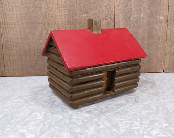 May include: A wooden log cabin toy with a red roof. The cabin has a small window and a chimney. The toy is made of wood and is painted brown and red.
