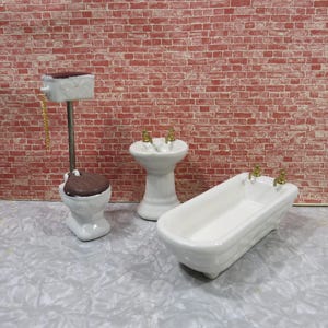 May include: A white porcelain dollhouse bathroom set with a toilet, sink, and bathtub. The fixtures have gold accents.
