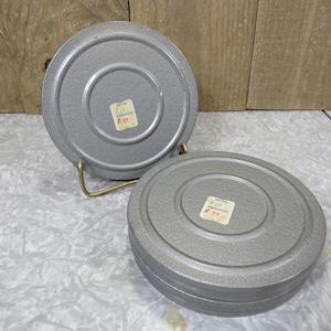 May include: Two vintage, gray metal film canisters with concentric circle detailing. One canister is open, revealing the interior, while the other is closed. Each has a small sticker with text. The canisters are on a gold-colored metal stand.